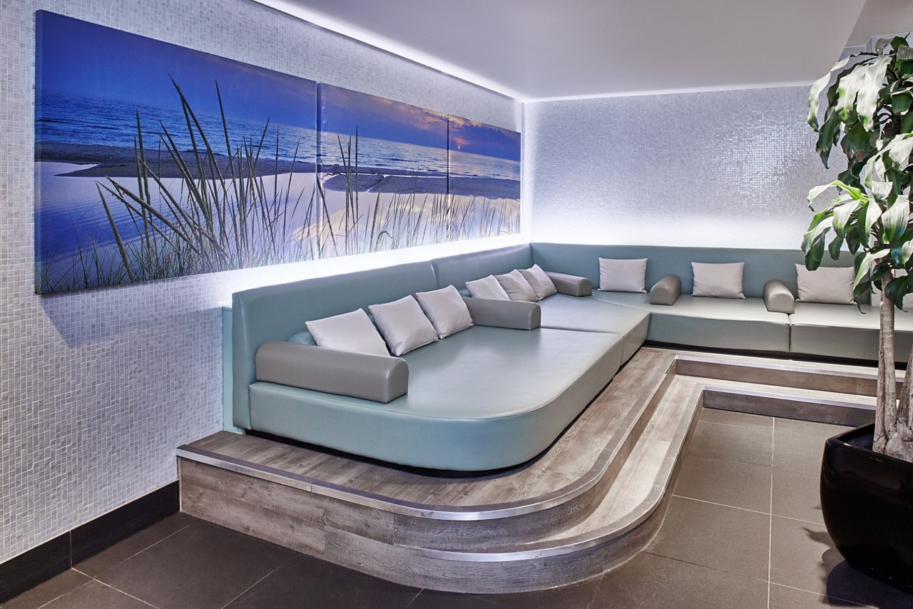 Curved sectional sofa extends along a corner, holding scattered gray pillows and bolsters; context: modern lounge with tiered wooden platform, mosaic-tile walls, beach triptych art, potted plant, and soft LED backlighting.